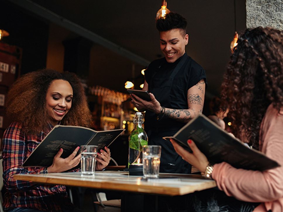 Two young women are at a trendy cafe ordering from restaurant menus that feature sustainability terms and phrases.